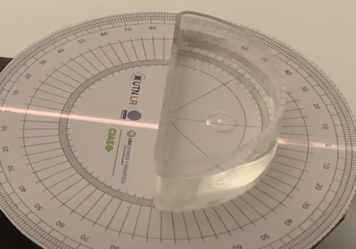 Vista superior real de uma célula semicircular com água sobre um transferidor impresso, atravessada por um raio laser vermelho.