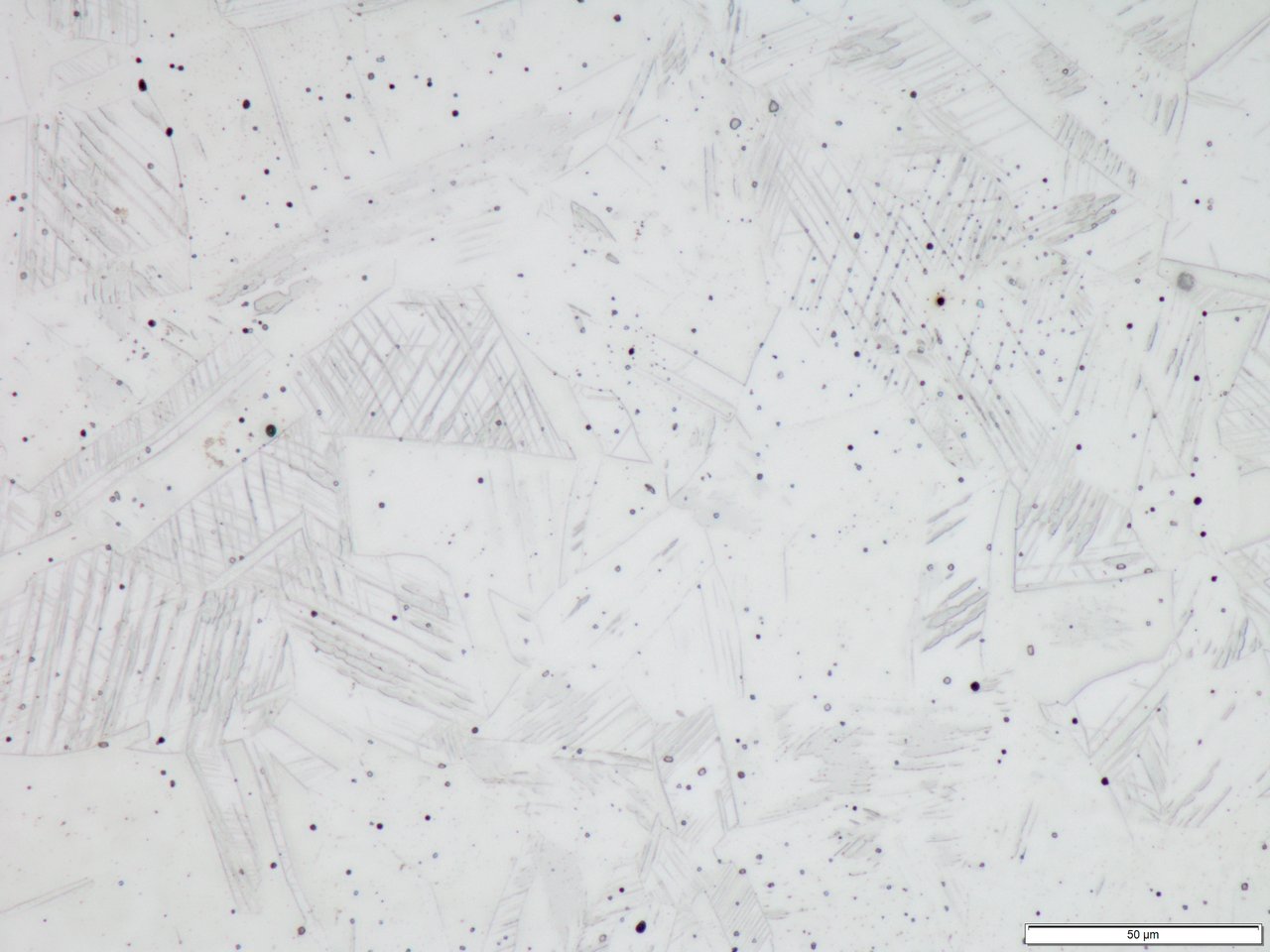 Micrografía asociada al acero 1.4301 con ataque químico a 50x, con regiones claras de contornos irregulares, varios grupos de líneas finas paralelas, puntos oscuros dispersos y una barra de escala de 50 µm.